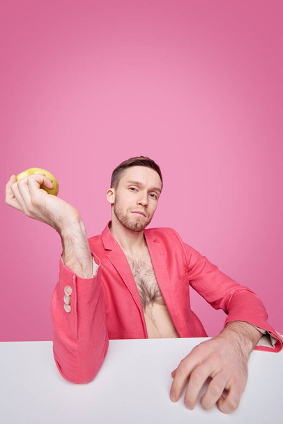 Jóképű fiatalember piros kabátban kezében alma vagy más gyümölcs, miközben pihentető íróasztal előtt kamera felett rózsaszín háttér - Fotó, kép