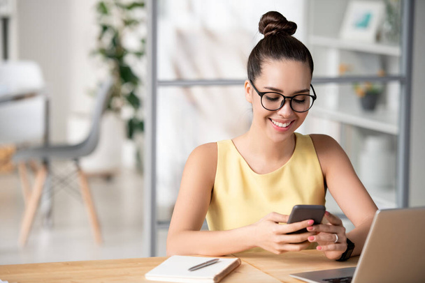 Νεαρή χαρούμενη γυναίκα διευθυντής κύλιση μέσα από τα μηνύματα στο smartphone, ενώ κάθεται μπροστά από το laptop και έχοντας διάλειμμα - Φωτογραφία, εικόνα
