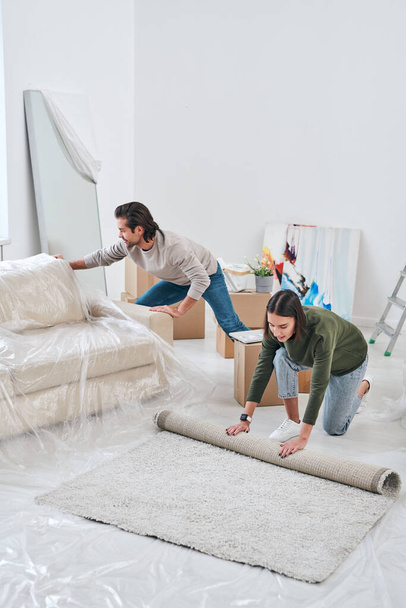 Young woman unrolling carpet on the floor of living-room while her husband removing new couch near by - Foto, Imagen