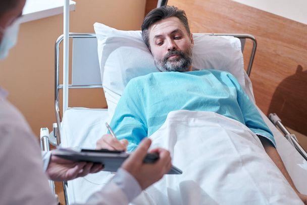 Volwassen bebaarde patiënt in ziekenhuisjurk liggend in bed en ondertekening van een medische overeenkomst terwijl hij toestemming geeft voor chirurgische ingreep - Foto, afbeelding