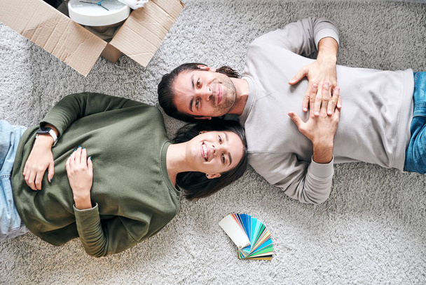 Vista general de la pareja joven alegre contemporánea en ropa de casualyacen en el suelo en su nueva casa o apartamento
 - Foto, imagen