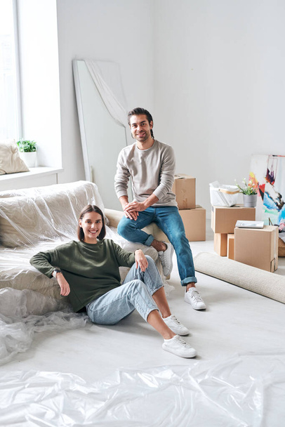 Young amorous couple in casualwear looking at you while relaxing by new couch in living-room of their flat or house - 写真・画像