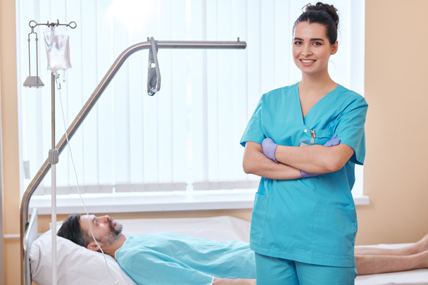 Portret uśmiechniętej młodej pielęgniarki w rękawiczkach chirurgicznych stojącej ze skrzyżowanymi ramionami przy łóżku pacjenta w szpitalu - Zdjęcie, obraz