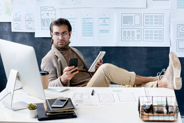 Pewny siebie mężczyzna w średnim wieku w okularach siedzący ze stopami na stole i odpowiadający na wiadomość telefoniczną analizując online szkic projektu aplikacji - Zdjęcie, obraz