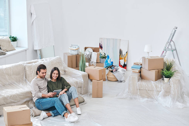Щаслива молода жінка і її чоловік сидять на килимі на дивані і прокручують через інтернет-магазин меблів
 - Фото, зображення