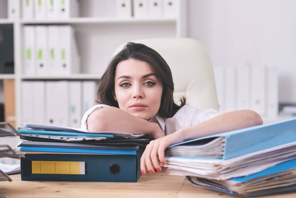 Portret van jonge zakenvrouw uitgeput met papierwerk leunend op mappen met werkdocumenten in kantoor - Foto, afbeelding