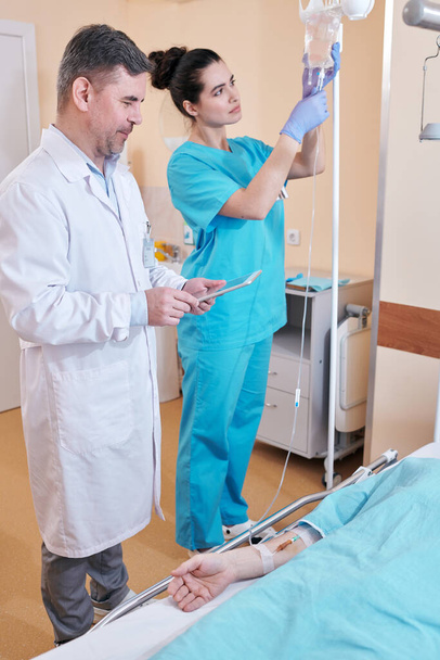 Jonge verpleegkundige bereidt infuuszak voor terwijl ze patiënt IV geeft, volwassen arts praat met patiënt terwijl ze zijn testresultaten bekijkt op tablet - Foto, afbeelding