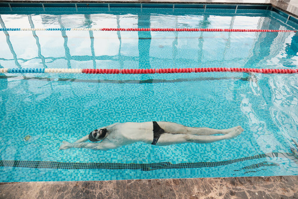 Above of young bearded man in goggles and cap swimming on back in modern pool - Foto, Imagem