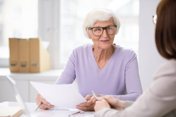 Старшая улыбающаяся клиентка со страховой формой, слушающая своего агента, объясняющего условия договора
 - Фото, изображение