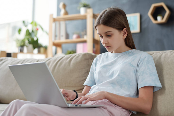 Gündelik giysili genç kız ev ortamında internette gezinirken dizlerinin üstünde kanepede oturuyor. - Fotoğraf, Görsel