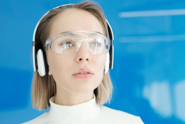 Primer plano de la joven pensativa en gafas innovadoras y auriculares inalámbricos mirando a su alrededor
 - Foto, Imagen
