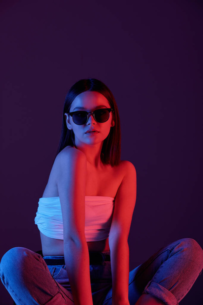 Молода стильна жінка в сонцезахисних окулярах, біла настільна і сині джинси, сидячи на підлозі студії на темному фоні
 - Фото, зображення