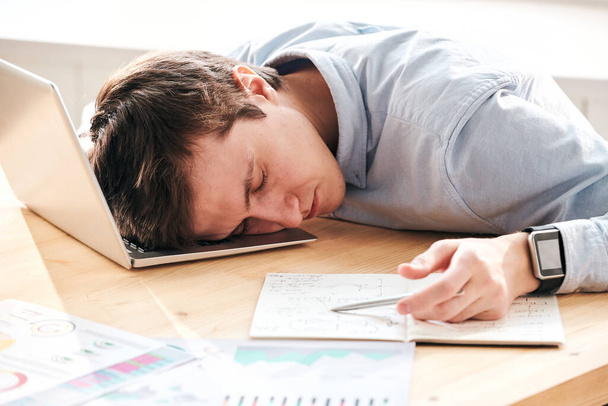 Joven hombre cansado agotado con cálculos acostado en el ordenador portátil mientras duerme en el lugar de trabajo
 - Foto, Imagen