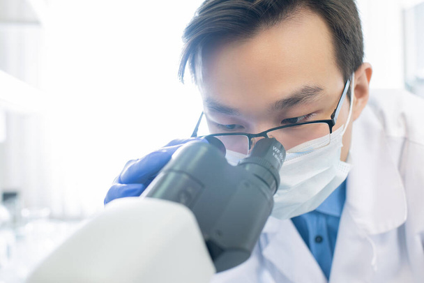 Scienziato di sesso maschile cinese in maschera e occhiali guardando al microscopio durante l'esperimento scientifico con nuovo vaccino in laboratorio
 - Foto, immagini