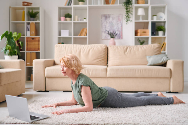 Mature jolie femme blonde en vêtements de sport couché devant un ordinateur portable tout en répétant l'exercice après l'instructeur pendant le cours de fitness en ligne
 - Photo, image