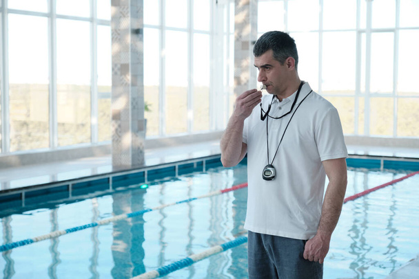 Experienced gray-haired male coach with stopwatch hanging on neck blowing whistle to swimmer in indoor pool - 写真・画像