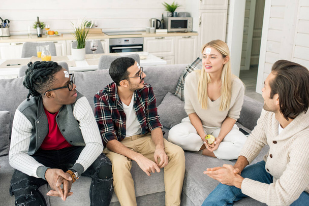 Čtyři mladí mezikulturní přátelé hrát název hry při relaxaci na gauči v obývacím pokoji při shromažďování doma - Fotografie, Obrázek