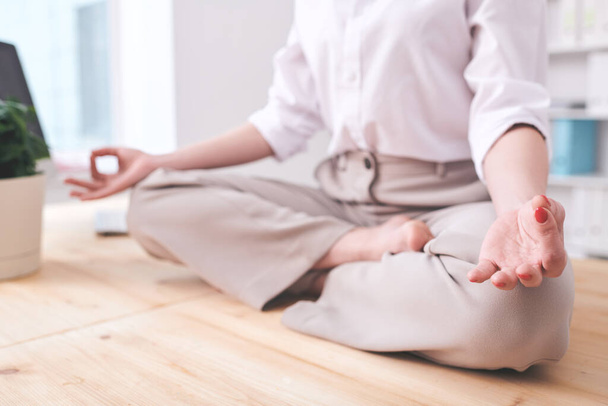 Zbliżenie nierozpoznawalnej bizneswoman siedzącej w pozycji lotosu i trzymającej się za ręce w mudrze podczas relaksującej medytacji w miejscu pracy - Zdjęcie, obraz