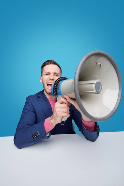 Enojado joven gerente masculino o jefe gritando en megáfono mientras que hace anuncio o advertencia contra fondo azul
 - Foto, imagen