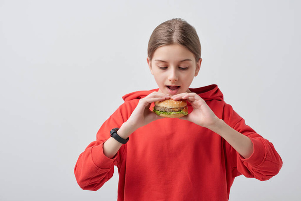 Nuorekas tyttö punainen huppari tilalla ruokahalua hampurilainen hänen avoin suu samalla syödä sitä valkoista taustaa vasten eristyksissä
 - Valokuva, kuva