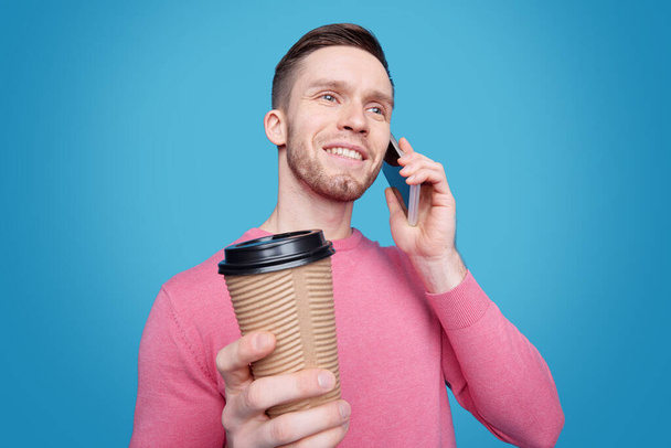 Feliz joven con sonrisa dentada hablando en el teléfono móvil mientras toma café frente a la cámara sobre fondo azul
 - Foto, Imagen
