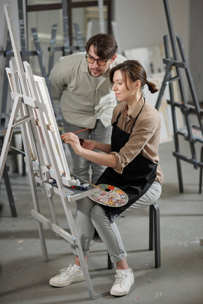 Красивая молодая студентка работает над живописью перед мольбертом, пока учитель наклоняется над ней во время консультации на уроке
 - Фото, изображение
