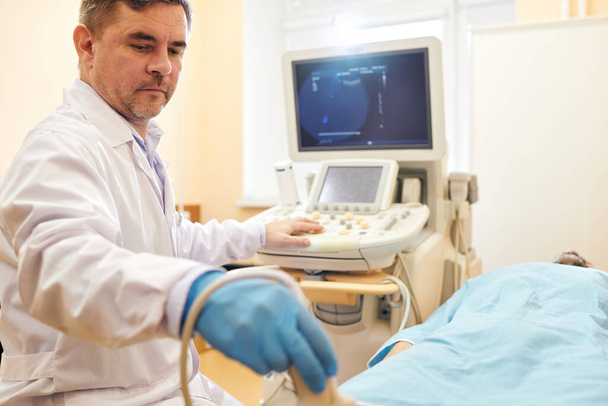 Poważny dojrzały lekarz w fartuchu laboratoryjnym stosujący sondę podczas wykonywania zabiegu ultrasonograficznego do pacjenta szpitalnego - Zdjęcie, obraz