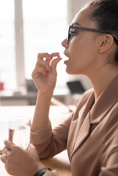 Jeune femme d'affaires élégante avec un verre d'eau va prendre la pilule ou de la vitamine tout en le tenant par la bouche
 - Photo, image