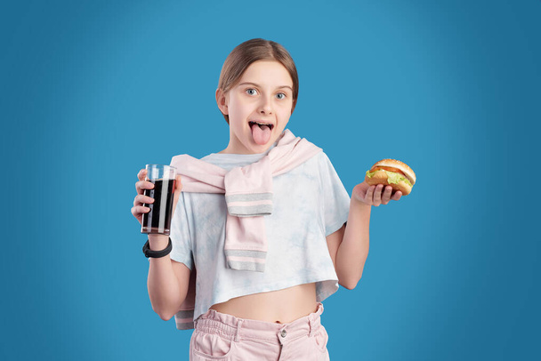 Zabawna nastolatka w stroju casualwear pokazuje Ci język podczas jedzenia hamburgera i picia coli na niebieskim tle - Zdjęcie, obraz