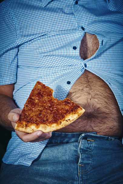 Nahaufnahme eines kaukasischen Mannes mit haarigem Bierbauch, der auf dem Sofa sitzt und Pizza isst - Foto, Bild