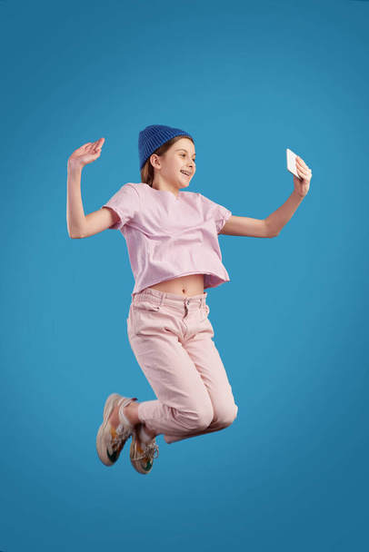 Екстатична дівчина-підліток в повсякденному одязі тримає смартфон перед собою, роблячи селфі в стрибку на синьому фоні
 - Фото, зображення