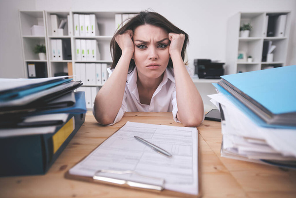 Portrait of frowning young businesswoman confused with paperwork sitting with head in hands at office table - Photo, Image