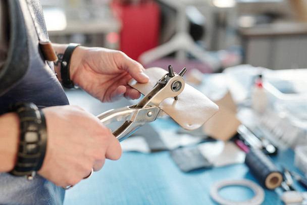 Mãos de artesão em avental com ferramenta especial fazendo furos em pequeno pedaço de couro bege enquanto está em pé perto do local de trabalho
 - Foto, Imagem