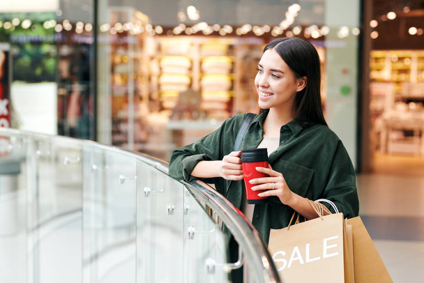 Lächelndes hübsches brünettes Mädchen steht am Geländer und trinkt heißen Tee aus Thermoskanne in der Halle des Einkaufszentrums - Foto, Bild