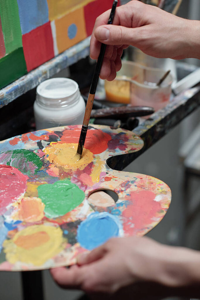 Main de jeune peintre professionnel mélangeant les couleurs sur la palette tout en se tenant devant le chevalet avec image inachevée
 - Photo, image