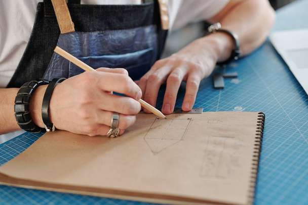 Χέρι του νέου τεχνίτη με μολύβι πάνω από τη σελίδα του σημειωματάριο, ενώ σχέδιο του αντικειμένου για να κάνει για έναν από τους πελάτες του - Φωτογραφία, εικόνα