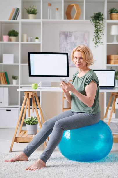 Довольно зрелая женщина со смартфоном сидит на фитбол во время поиска онлайн фитнес-курс во время самоизоляции дома
 - Фото, изображение