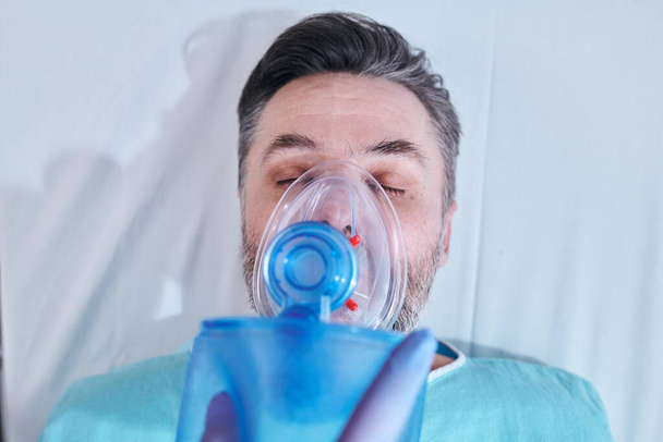 Edellä näkymä kypsä potilas vakavassa kunnossa pitää silmät kiinni hengittäessään happea pussi venttiili naamio
 - Valokuva, kuva