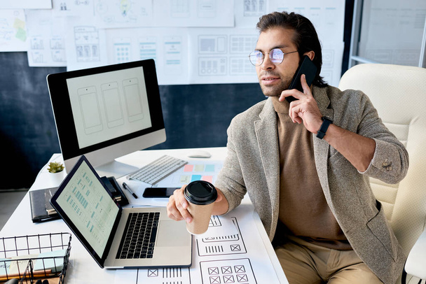 Moderní sebevědomý muž manažer v brýlích sedí za stolem a pije kávu, zatímco mluví se zákazníkem o designu aplikace - Fotografie, Obrázek