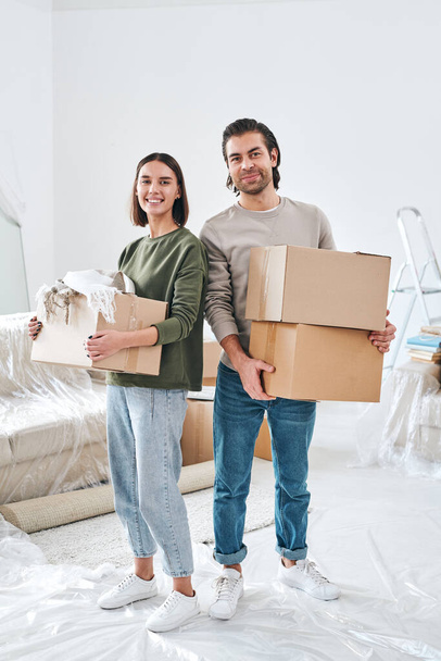 Veselý mladý pár v neformálním oblečení držící kartónové krabice s balenými věcmi v obývacím pokoji svého nového domu - Fotografie, Obrázek