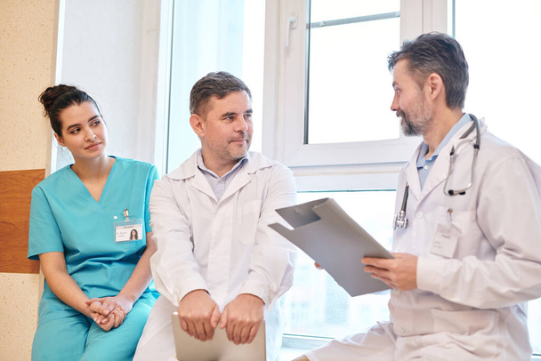 Бородатый врач со стетоскопом вокруг шеи рассказывает коллеге о новых лекарствах для пациента в коридоре больницы
 - Фото, изображение