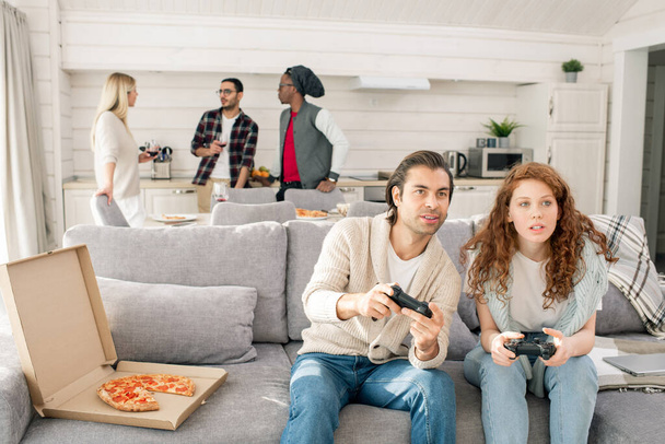 Giovane coppia con joystick seduti sul divano davanti alla TV e giocare ai videogiochi sullo sfondo dei loro amici
 - Foto, immagini
