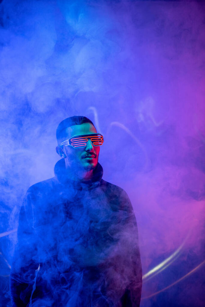 Plan studio d'un jeune homme barbu aux lunettes rouges en spirale debout dans la fumée au milieu de la lumière bleue au néon
 - Photo, image