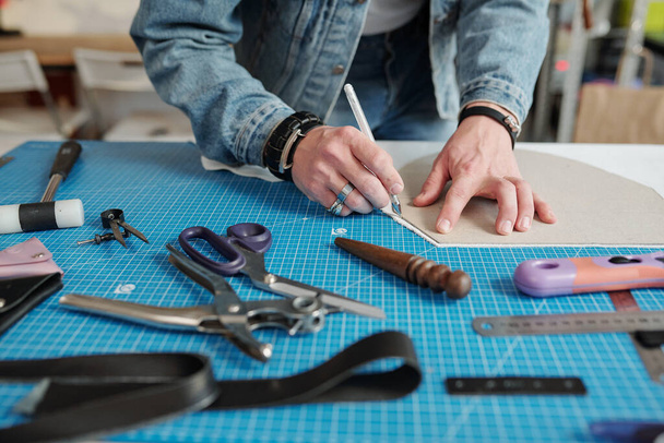 Manos de artesano joven inclinándose sobre la mesa mientras delinea el patrón de papel con la pluma entre los suministros de trabajo
 - Foto, imagen