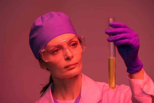 Młoda pracownica laboratorium w białym płaszczu, rękawiczkach ochronnych i okularach, patrząca na żółtą substancję płynną w kolbie - Zdjęcie, obraz