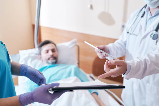 Крупный план неузнаваемой медсестры в перчатках, показывающей карточку пациента в блокноте врачу в больнице во время обсуждения лечения пациента - Фото, изображение