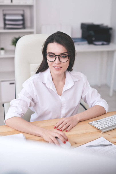 Χαμογελαστή νεαρή έξυπνη κυρία με γυαλιά που κάθεται στο γραφείο και χρησιμοποιεί σύγχρονο υπολογιστή στο γραφείο - Φωτογραφία, εικόνα