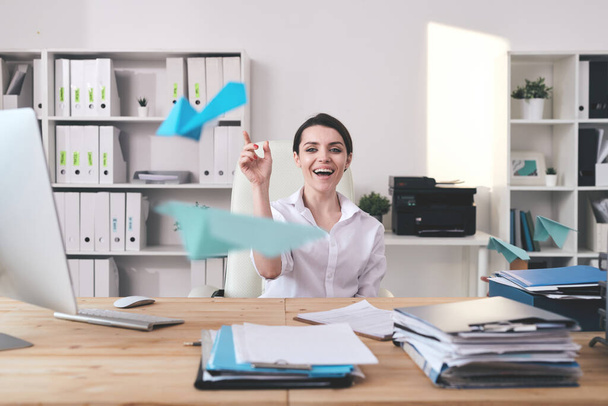 Cheerful excited young businesswoman sitting at desk full of documents and throwing paper plane in office - Foto, Imagem