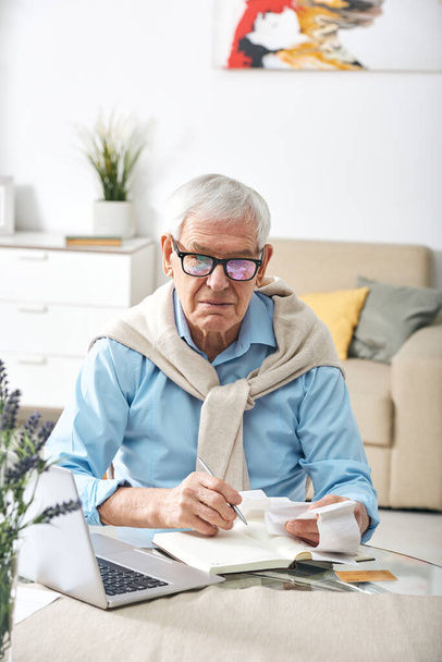 Egy komoly, modern, szemüveges idős férfi portréja, aki az asztalnál ül és a költségek kiszámítása során ellenőrzi az ellenőrzéseket. - Fotó, kép