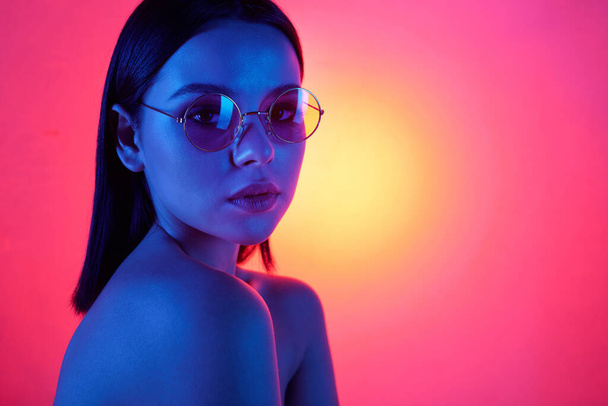 Young brunette female in stylish eyeglasses with perfect smooth hair and natural makeup looking at you on neon pink background - Фото, зображення
